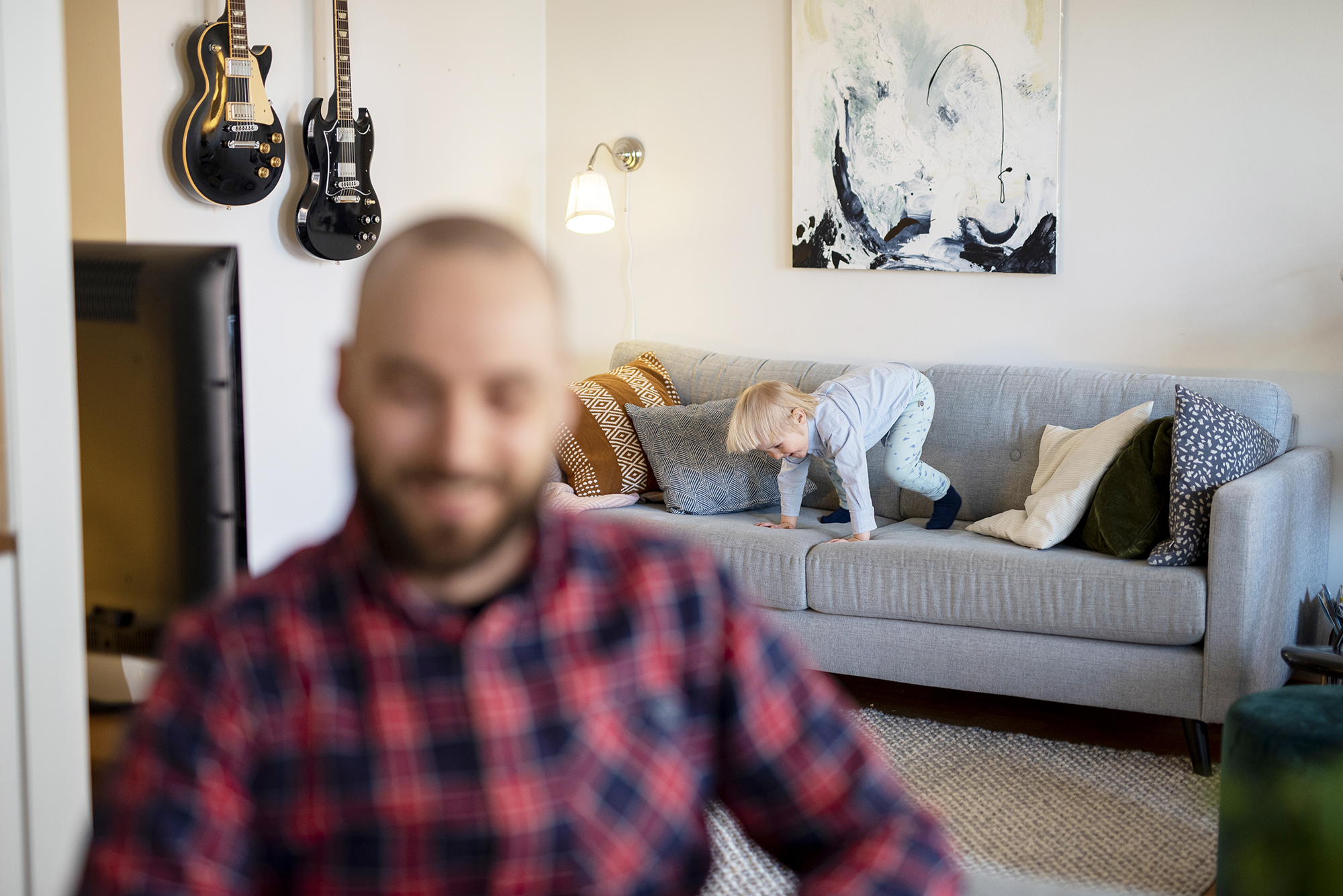 Mies hymyilee, taustalla lapsi tekee kuperkeikkaa sohvalla